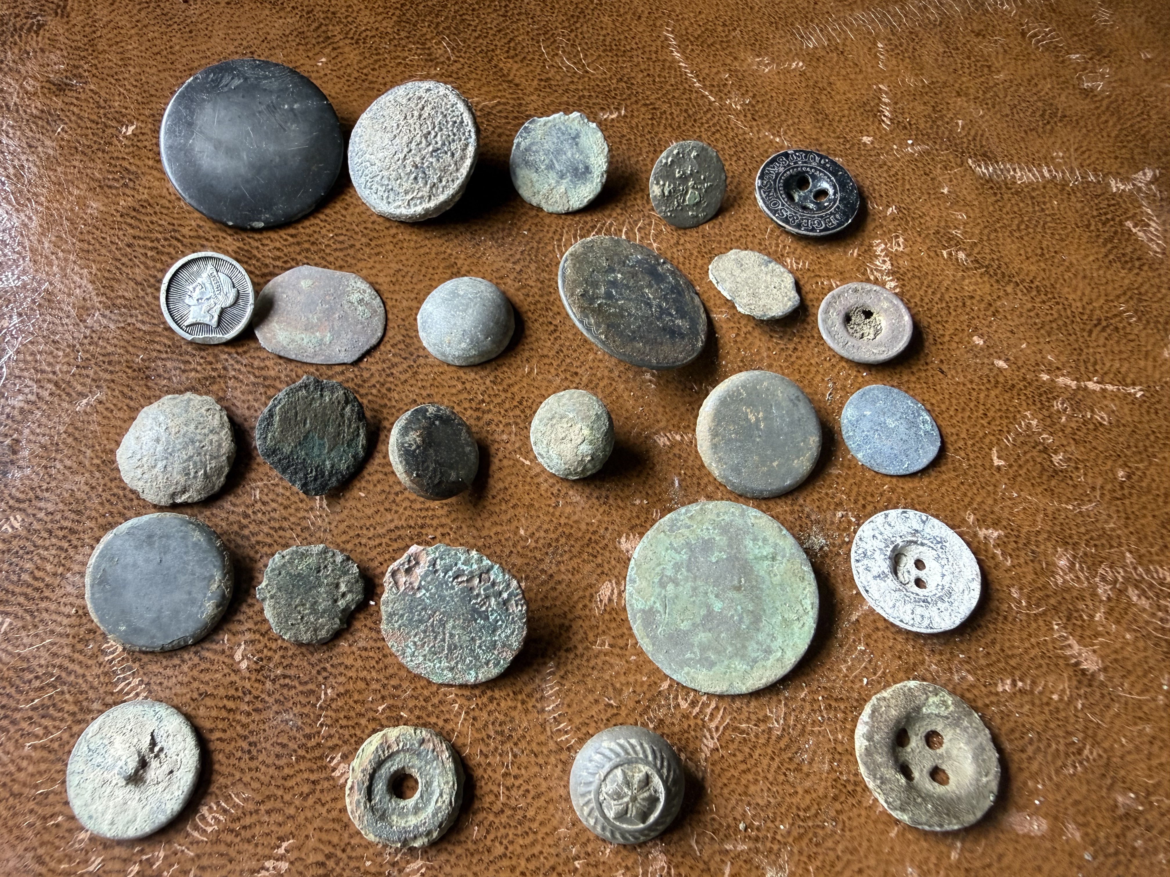 Large collection of corroded buttons, coins, and discs of varying sizes arranged on brown leather, found by metal detecting
